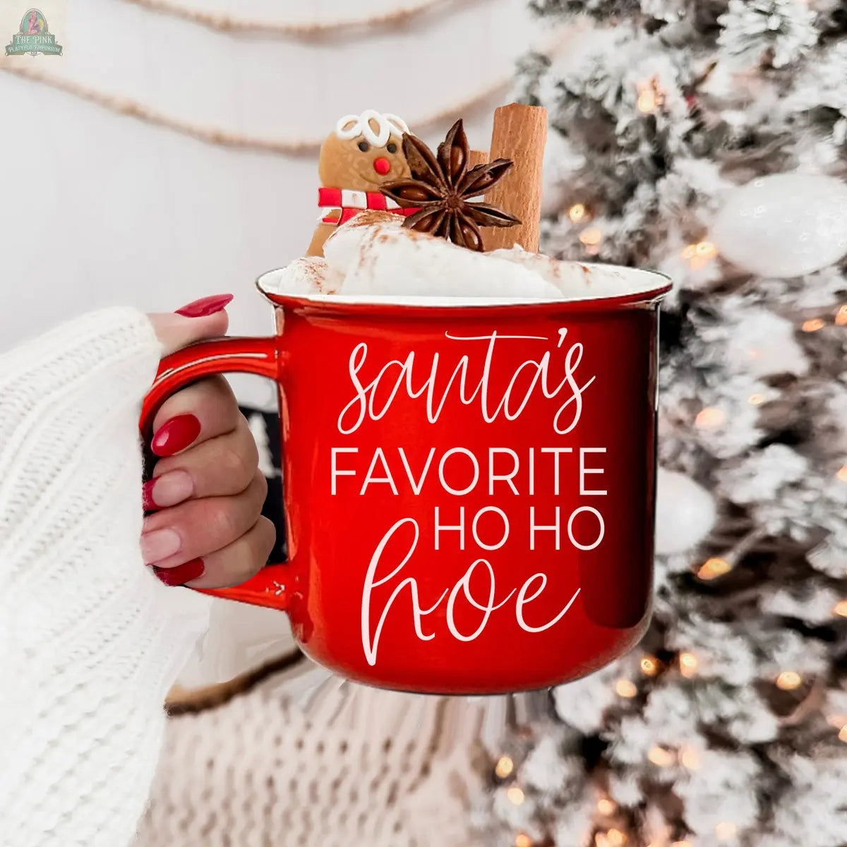 A hand with red nails holds the Ho Ho Hoe Mug, filled with whipped cream, a gingerbread cookie, cinnamon stick, and star anise. The ceramic, microwave-safe mug features “Santa’s favorite ho ho hoe.” A glowing, snowy Christmas tree is in the background.