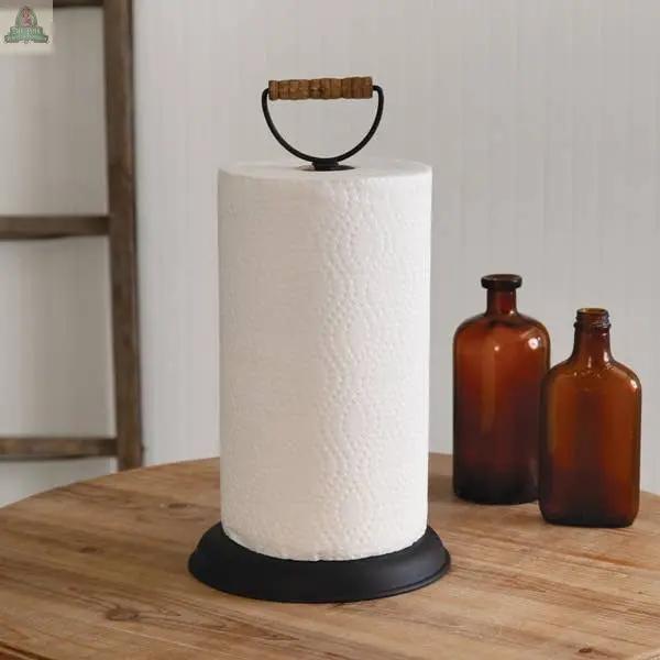The Homestead Paper Towel Holder, featuring a black metal base and wooden handle, is displayed on a wooden table beside two brown glass bottles.