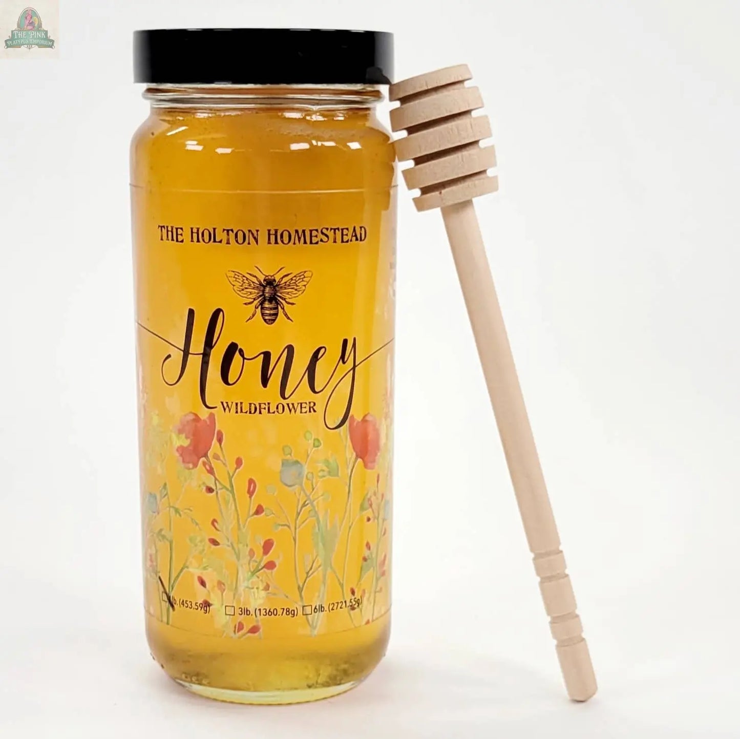 A Pink Platypus Emporium Wood Honey Dipper, an eco-friendly kitchen gadget, is paired with a glass jar of wildflower honey labeled “The Holton Homestead Honey” featuring bee and floral designs on a white background.