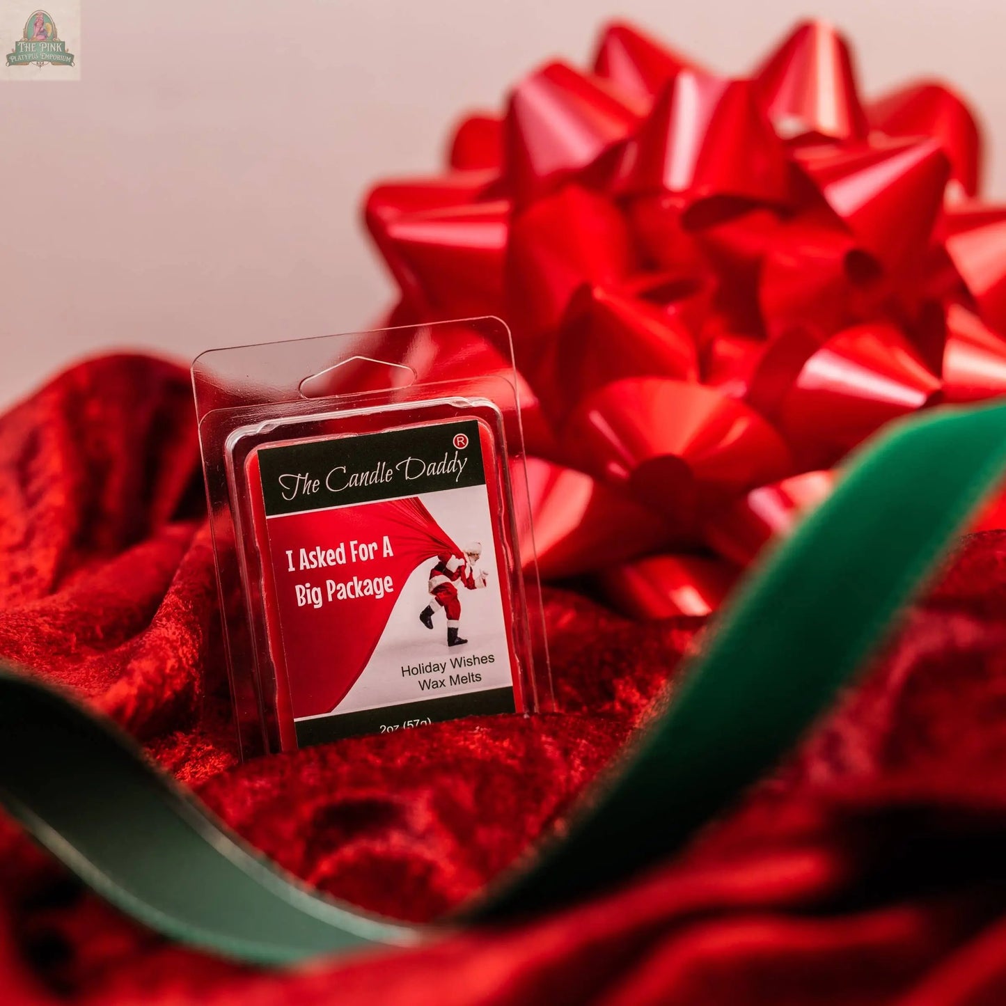 A pack of I ASKED FOR A BIG PACKAGE - HOLIDAY WISHES SCENTED WAX MELTS sits on red velvet with a big red bow and green ribbon in the background—ideal for bringing festive aromatherapy to your holiday décor.