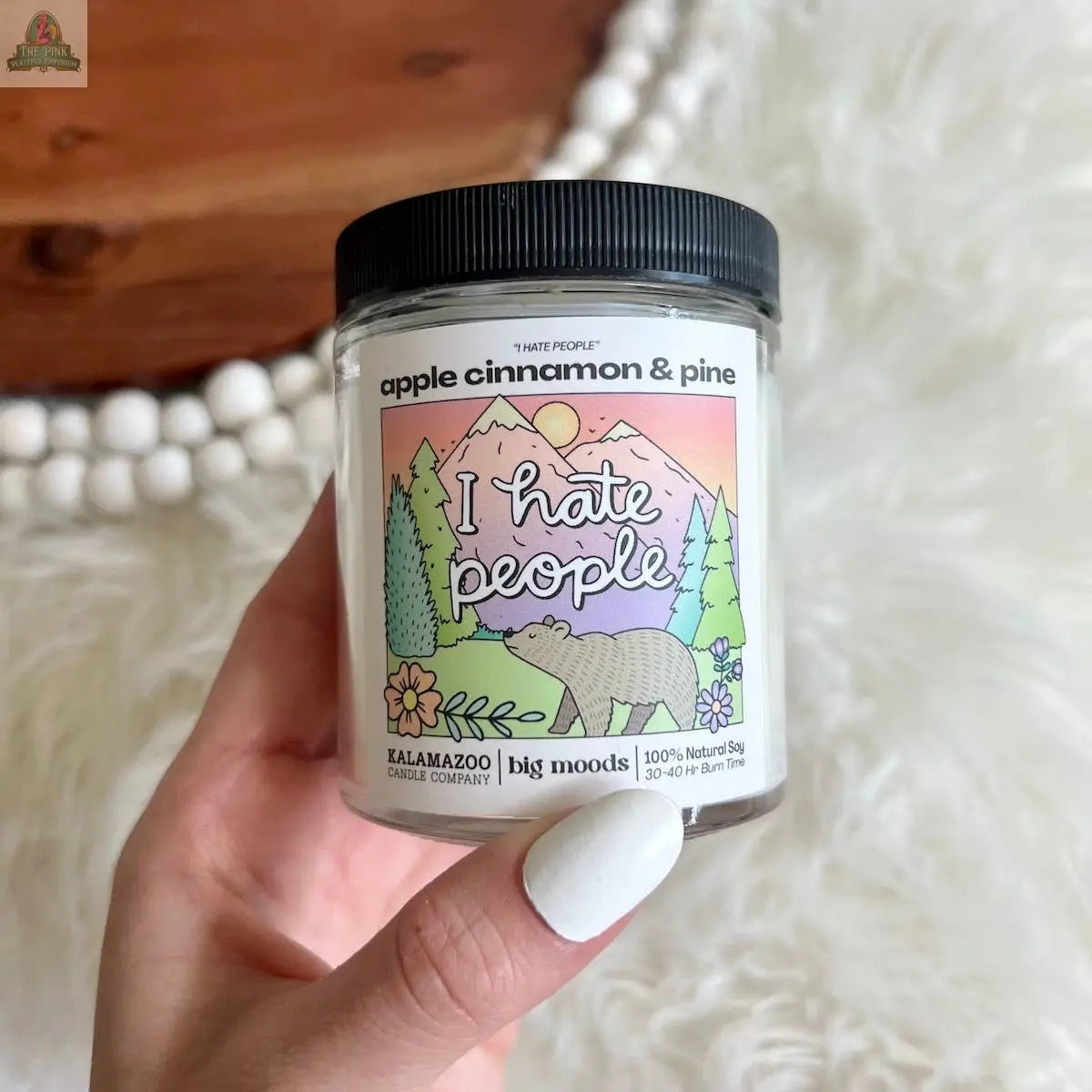 A hand with white-painted nails holds a "I Hate People" Apple Cinnamon & Pine Luxury Soy Candle by Pink Platypus Emporium, featuring a label with a bear, trees, and mountains.