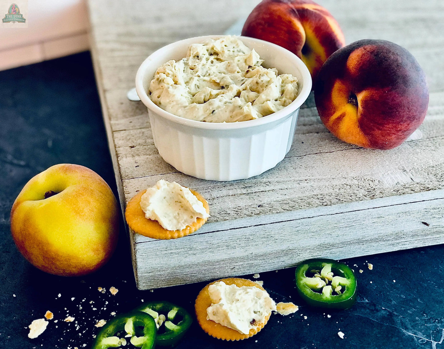 A white ramekin with Carmie's Kitchen Jalapeno Peach Dip Mix sits on a wooden board, surrounded by peaches, jalapeños, and crackers topped with the creamy dip—ideal for serving or gifting at your next holiday party.