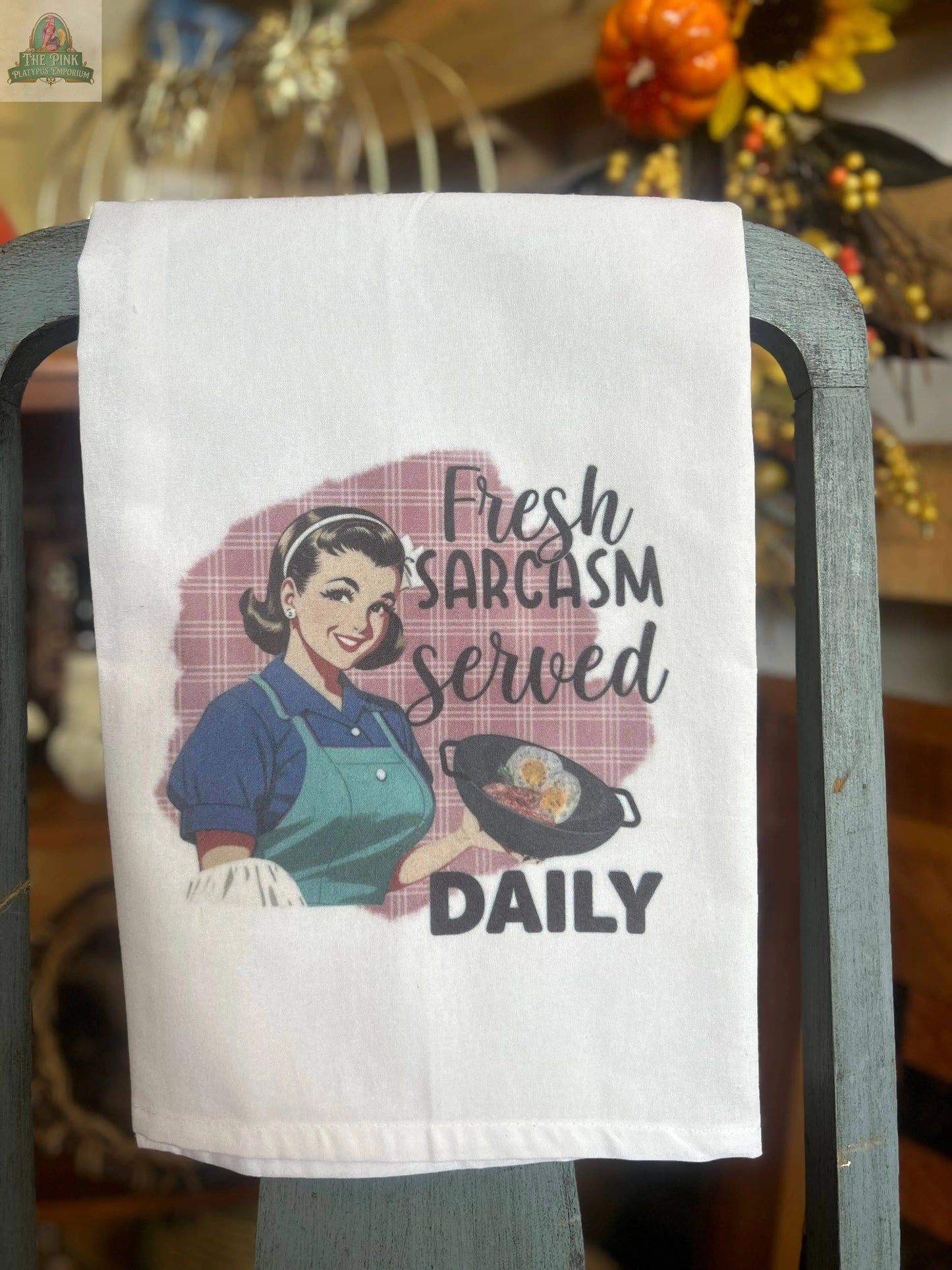 A Pink Platypus Emporium Funny Tea Towel hangs on a chair, featuring a retro woman with a frying pan and the text "fresh sarcasm served daily." Softly blurred autumn decor appears in the background.