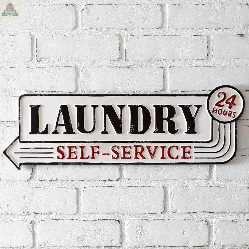 The Laundry Metal Sign is a wall mount metal sign that reads "Laundry Self-Service" with "24 Hours" inside a red circle, black and red text, a left-pointing arrow, and keyhole picture hangers for easy hanging on your white brick wall.