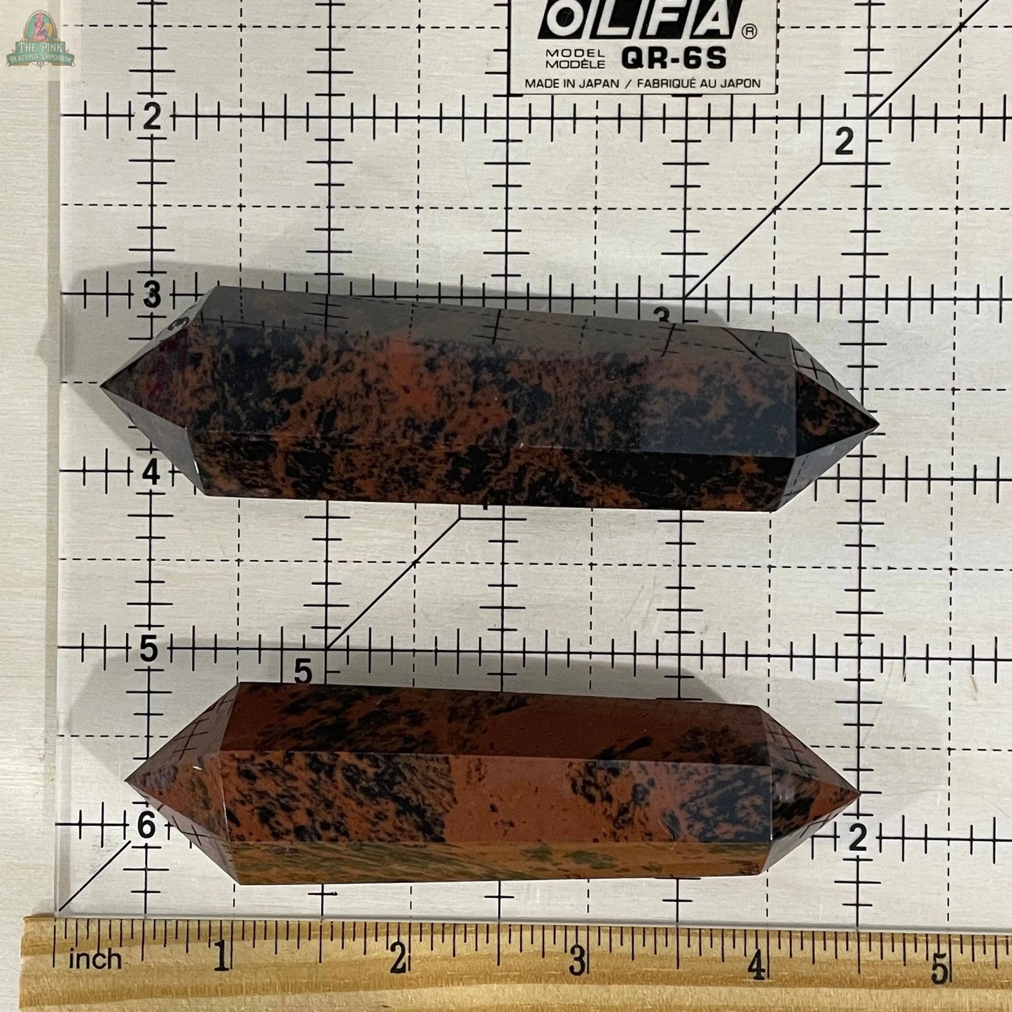 Two Keystone Crystals Mahogany Obsidian Double Terminated 3 1/2" Obelisks, featuring reddish-brown and black tones, are displayed horizontally on a cutting mat grid with ruler for scale.