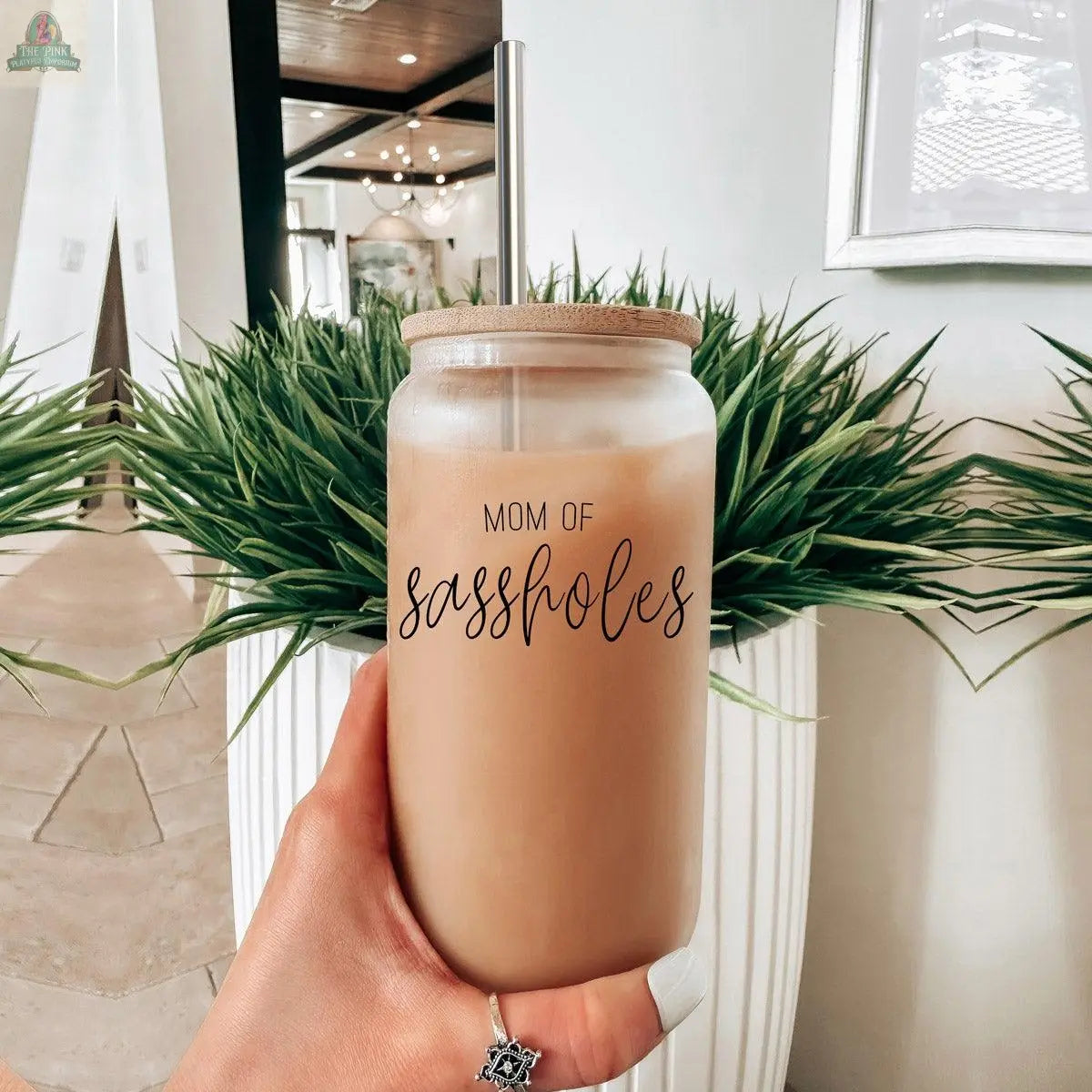 A hand wearing a ring holds the 17oz "Mom of Sassholes" glass iced coffee jar with bamboo lid and stainless steel straw; green plants and modern decor are in the background.