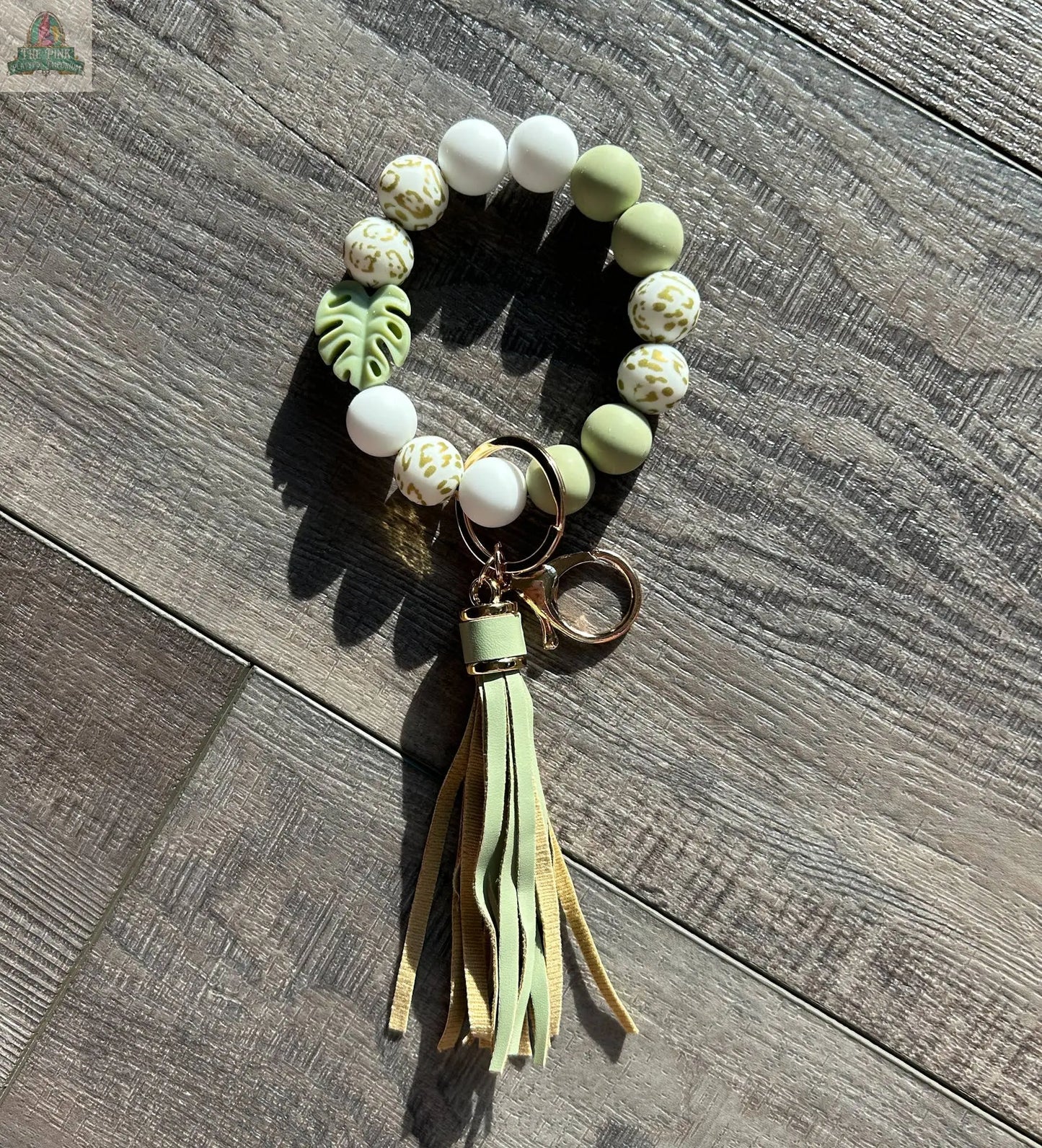 The Monstera Leaf Lover Beaded Wristlet Keychain by Pink Platypus Emporium, featuring white, green, and patterned beads, a green Monstera leaf bead, gold key ring, and pale green tassel—perfect for plant lovers—rests on a wooden surface.