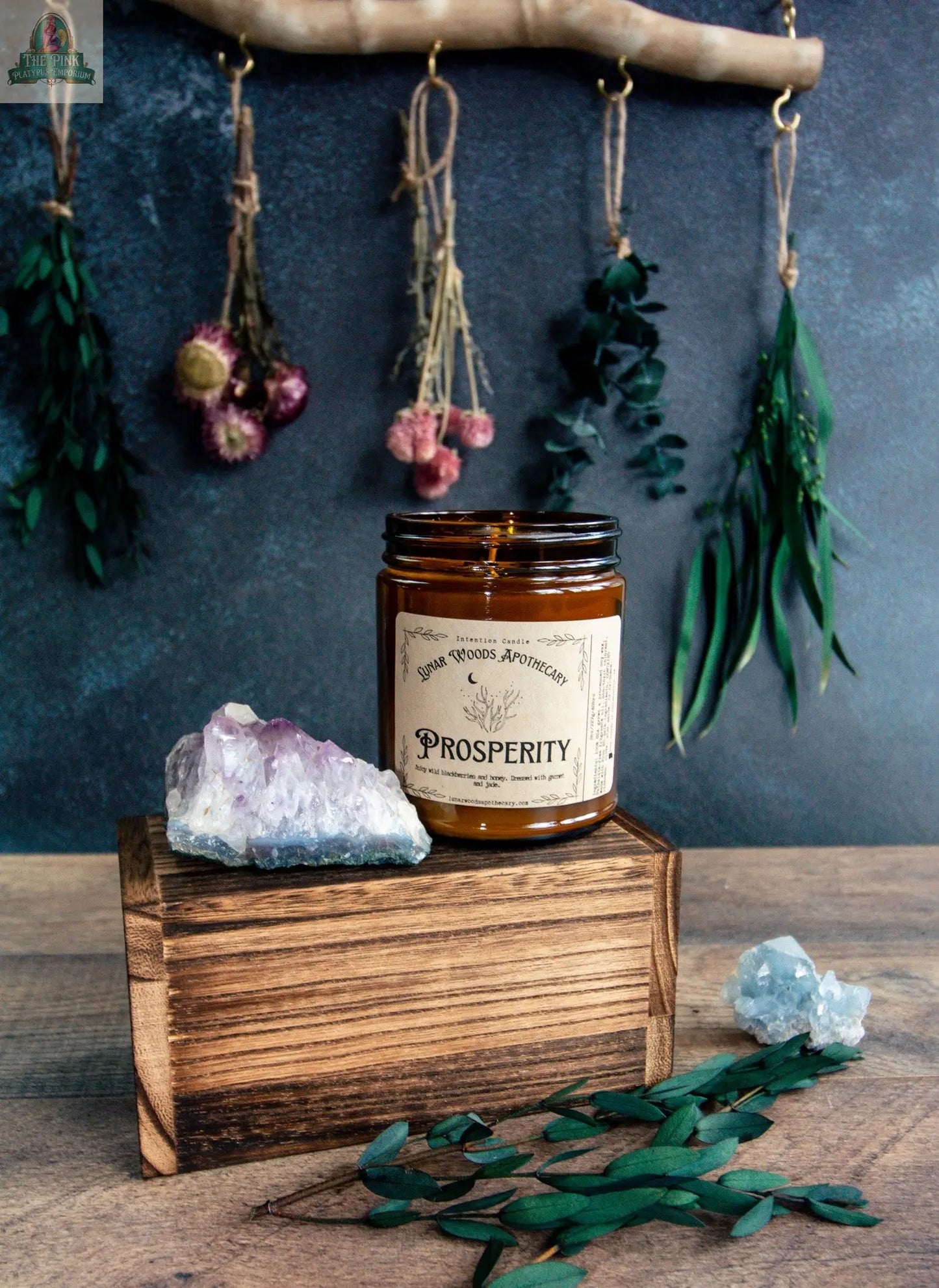 A brown glass NEW Prosperity candle sits on a wooden box beside crystals and green leaves. In the background, dried flowers and herbs hang from a wooden branch against a dark wall, creating an abundant atmosphere.