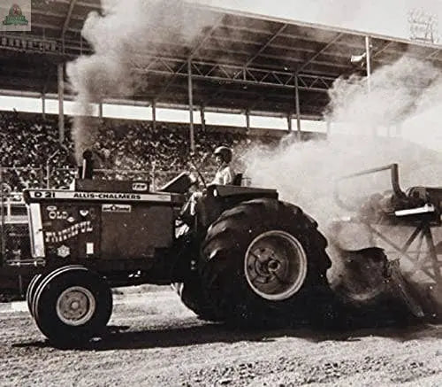 At a tractor pull event, thick smoke rises as someone drives an Allis-Chalmers tractor. Among the crowd, one fan stands out in a Pink Platypus Emporium Funny BBQ Apron—“PULL MY PORK”—featuring handy beer pockets. Black and white photo.