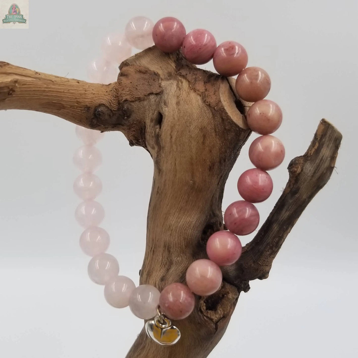 The Pink Platypus Emporium Natural Rose Quartz Stone Heart Bracelet, featuring alternating pink beads and a heart charm, is elegantly draped over driftwood for display against a clean white background.