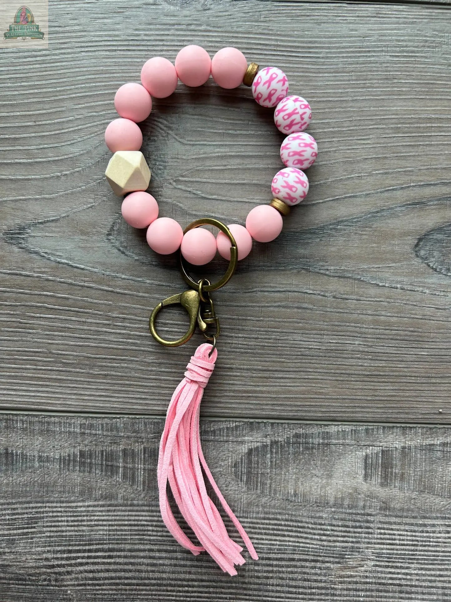 The Breast Cancer Pink Ribbon Beaded Wristlet by Pink Platypus Emporium features pink beads, a gold clasp, suede tassel, and leopard print accents—perfect as a stylish breast cancer awareness accessory. Displayed on a wooden surface.