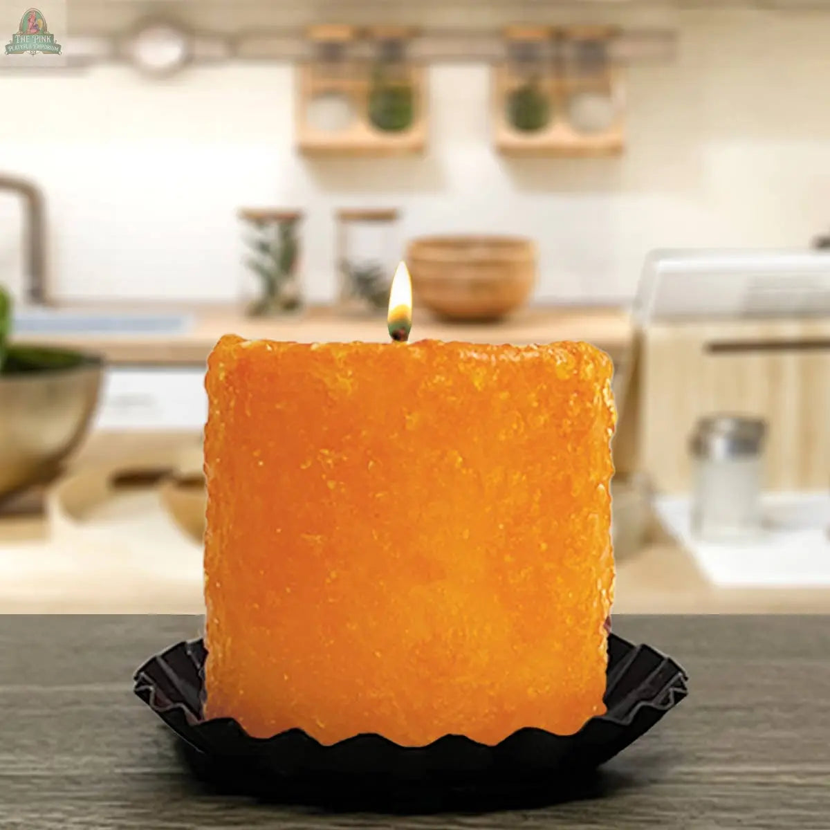 The Pink Platypus Emporium Orange Cranberry Mini Hearth, a square orange candle with a lit flame, sits on a black tray atop a dark surface, with a blurred kitchen background.