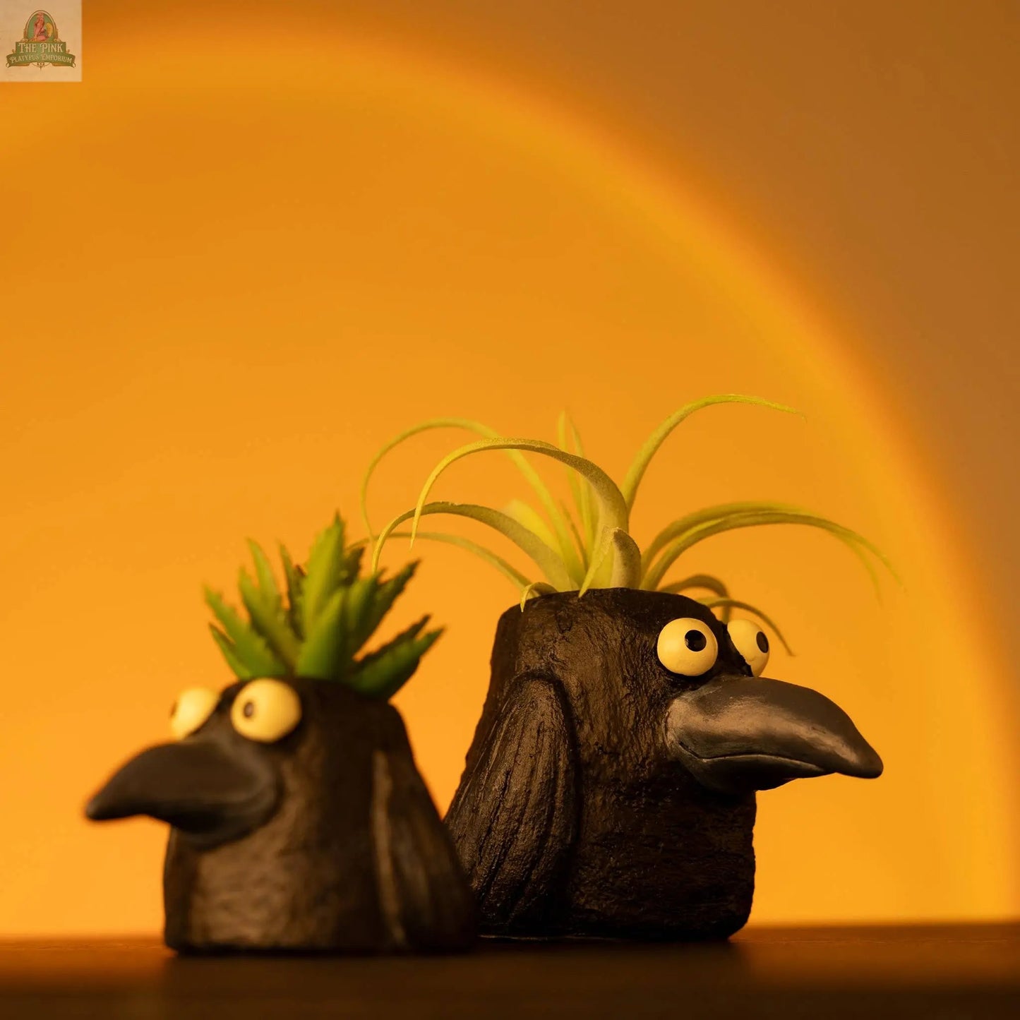 Two Poe the Crow Blobhouse Planters, featuring large-eyed black crow designs holding small green plants as "hair," are set against a warm yellow-orange gradient—ideal for adding a unique touch to your home décor.