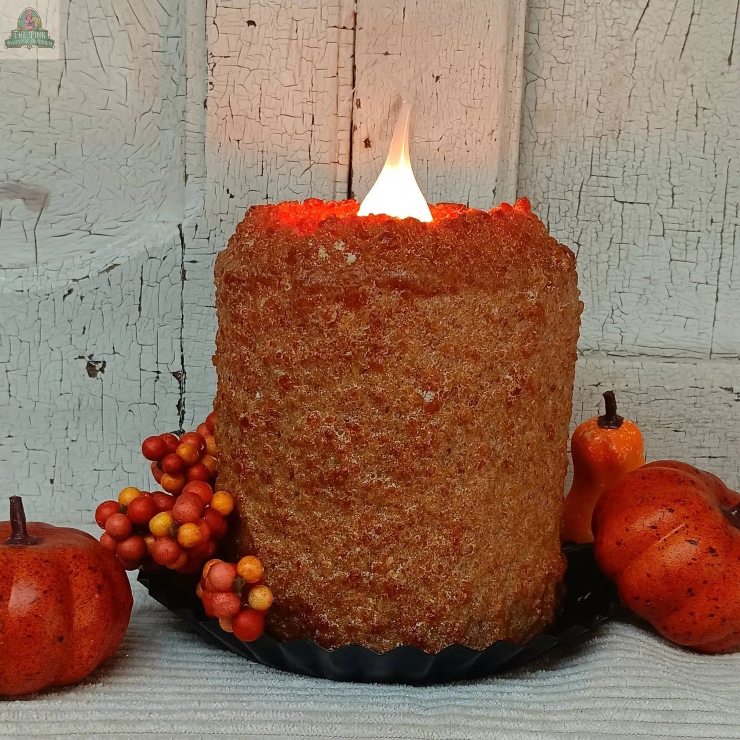 The Pumpkin Crumb Cake Electric Candle with a rustic orange texture sits on a black plate, surrounded by artificial autumn berries and mini pumpkins, set against a weathered white wooden background.
