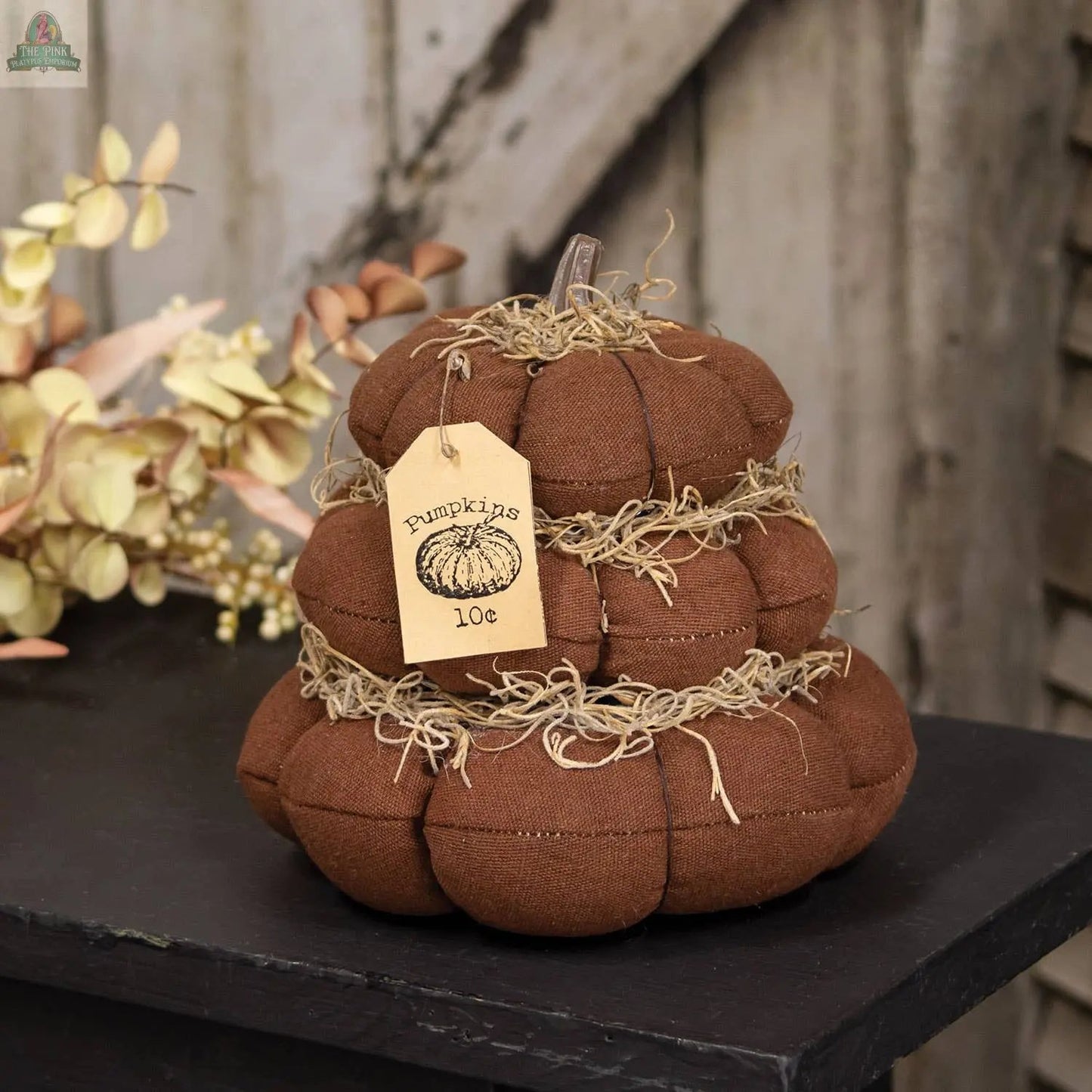 The "Pumpkins 10 Cents" Stuffed Pumpkin Stack features three brown plush pumpkins with straw accents and a tag, making it a charming rustic decoration for fall displays.