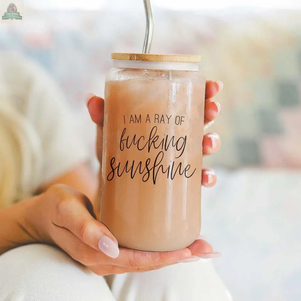 A person holds the Ray of Sunshine 17oz glass cup with a bamboo lid and stainless steel straw, filled with a light brown drink. The jar is printed with the phrase, "I am a ray of fucking sunshine.