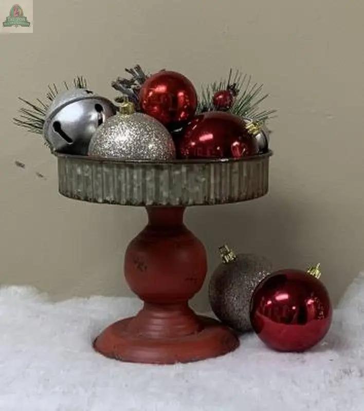 The Red Base Candle Pan displays red, silver, and glittery Christmas ornaments with pine sprigs; two ornaments sit on faux snow beside the pan, all arranged in front of a beige wall.