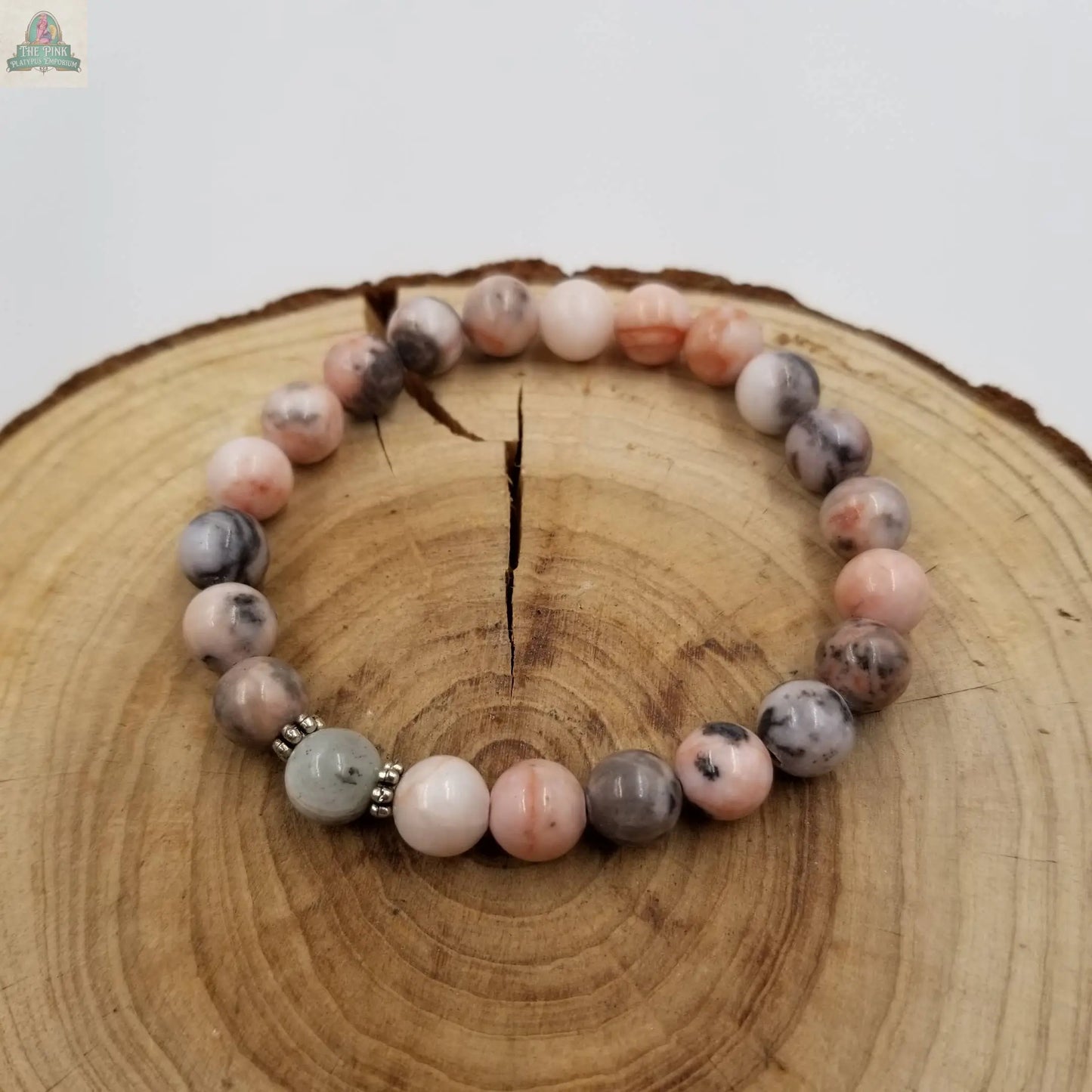 The Rhodonite & Labradorite Gift for Mom's Bracelet from Pink Platypus Emporium features polished pink, gray, and light green beads on a rustic wood slice—a stylish piece of healing crystal jewelry.