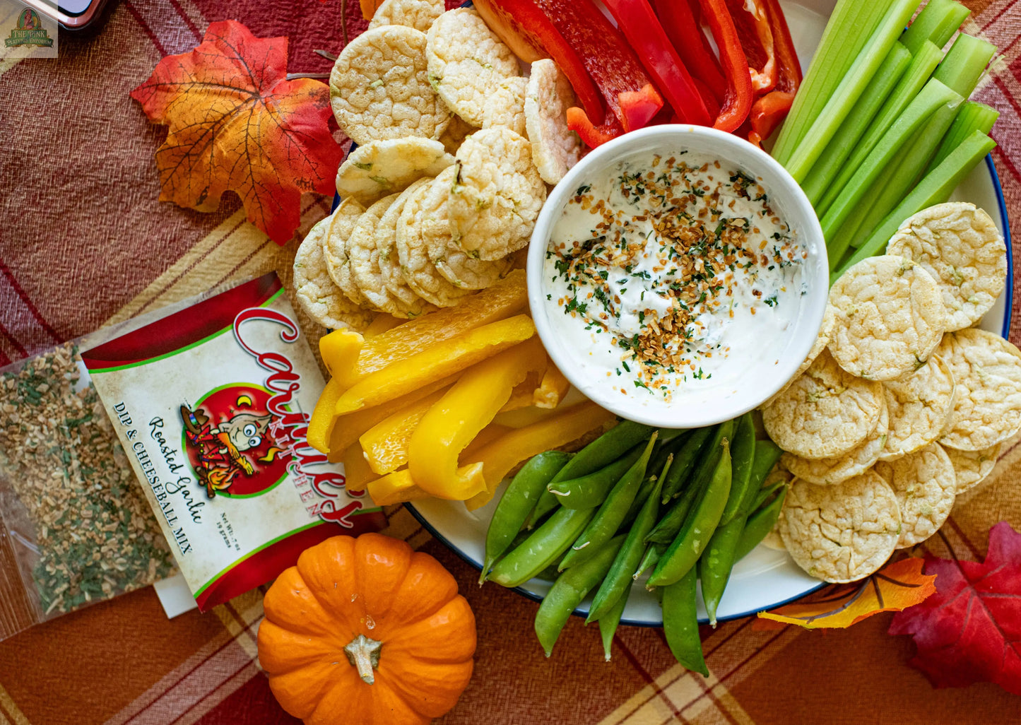 A festive platter of rice cakes, celery, bell pepper strips, and snap peas with a bowl of Carmie's Kitchen Roasted Garlic Dip Mix sits on an autumn tablecloth beside a mini pumpkin and the dip mix packet.
