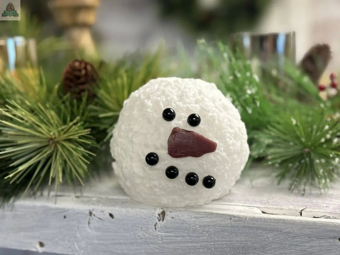 The Round Snowman Moving Flame LED Candle (4x3.75in) features a textured snowman face with a brown cone nose, black bead details, and is set against a background of pine branches and pinecones—perfect for charming wholesale home décor.
