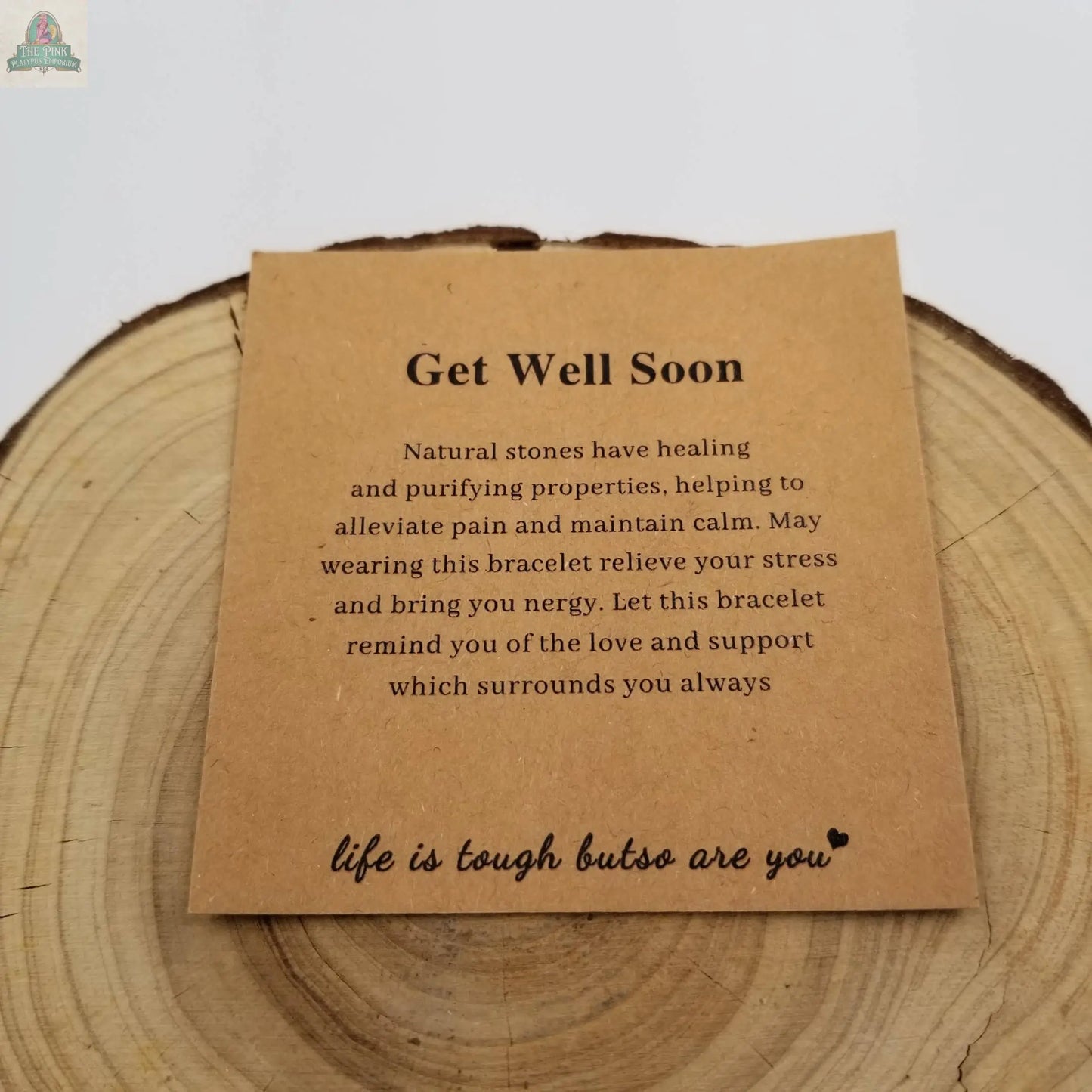 A brown card on wood reads: Get Well Soon. Pink Platypus Emporium’s Safe & Healthy Natural Stone Blessing Bracelet supports your Throat Chakra, purifies energy, and soothes pain—life is tough but so are you ♥.