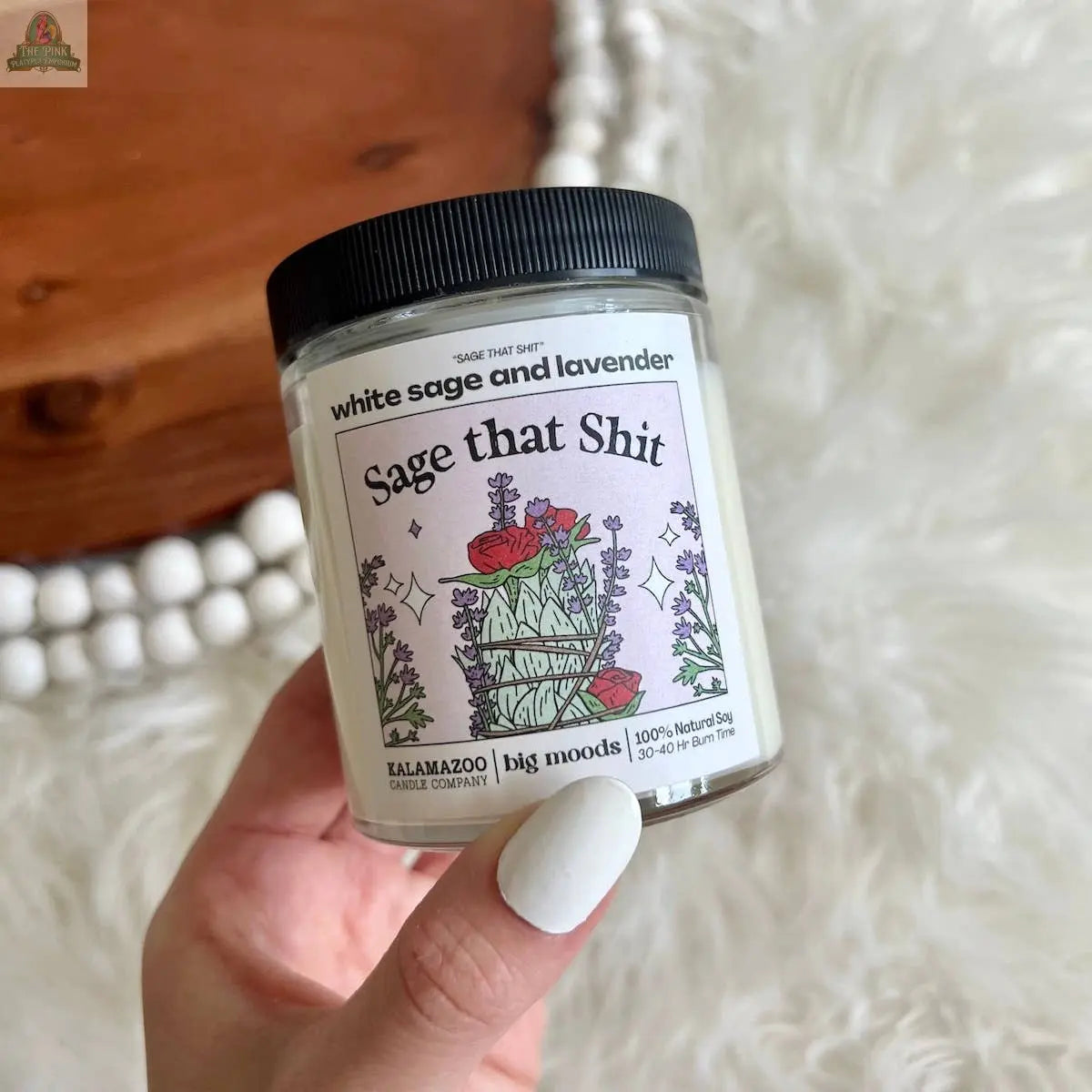 A hand with white nail polish holds Pink Platypus Emporium's "Sage That Shit" White Sage & Lavender Luxury Soy Candle, featuring sage, lavender, and crystal illustrations. In the background lies a white beaded garland on soft fabric.