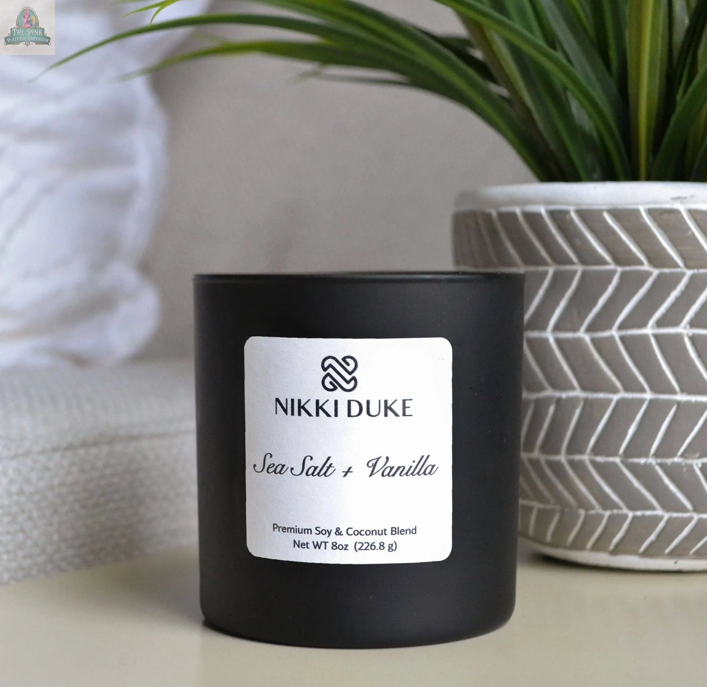 A black Sea Salt + Vanilla Premium Soy Candle by Pink Platypus Emporium sits beside a white textured plant pot with green leaves, creating a cozy ambiance reminiscent of an ocean paradise.