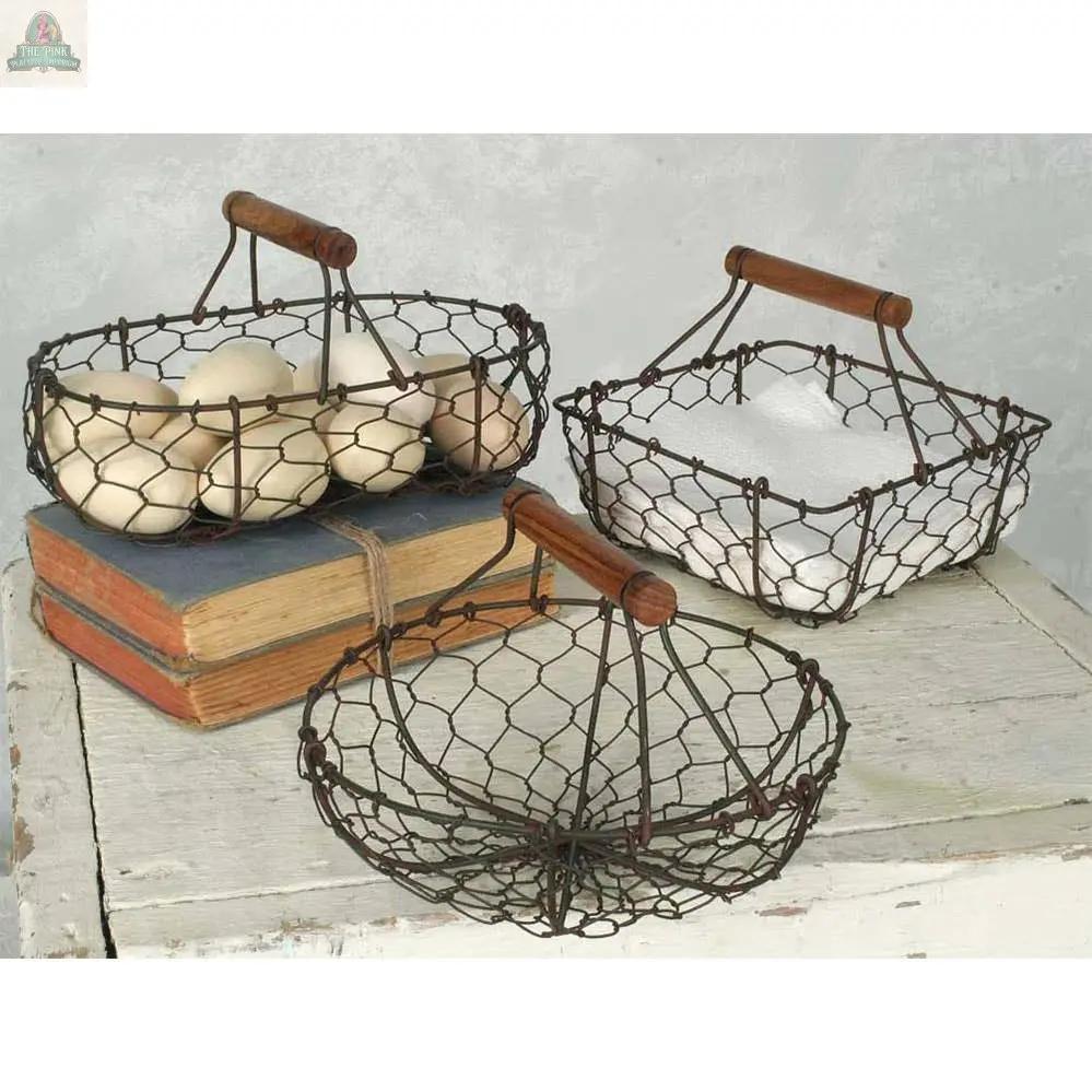 Three CTW Home Collection Chicken Wire Baskets—square, round, and oval with wooden handles—rest on a rustic white surface: one holds eggs, one is empty, and one displays a folded white cloth on two stacked vintage books.