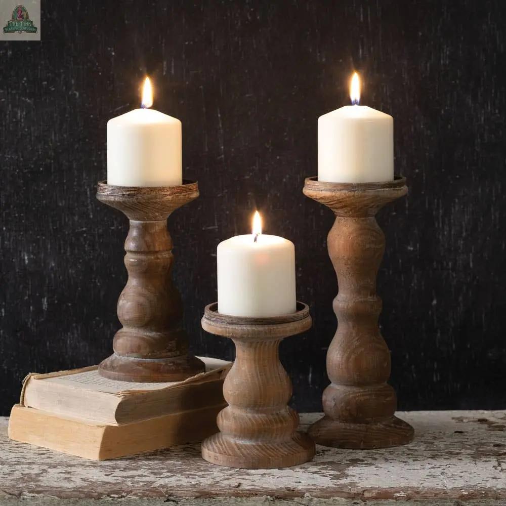 The Set of Three Wooden Pillar Candle Holders, displayed with white candles and one holder on a stack of vintage books, adds warm rustic charm against a dark textured background.