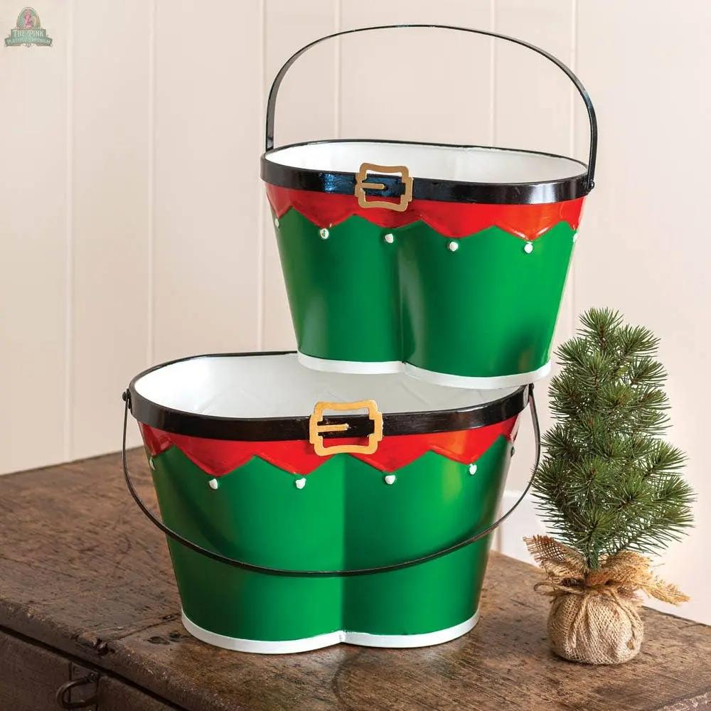 The Set of Two Elf Pants Buckets, featuring green buckets with red, black, and gold trim, are stacked on wood beside a small burlap-wrapped artificial pine tree, creating festive storage or decorative holiday display.