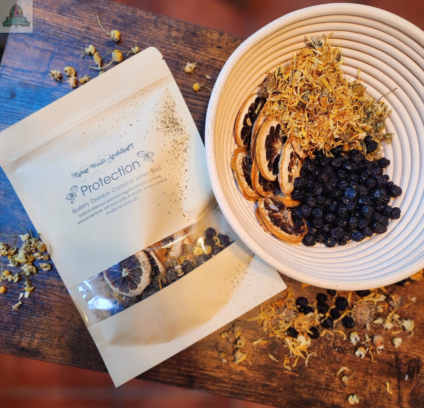 A bowl with dried blueberries, lemon slices, and herbs sits beside a kraft package labeled "Simmer Pot Kit- Protection" on a rustic wooden surface. Loose herbs are scattered nearby, all ready to help cleanse your space.