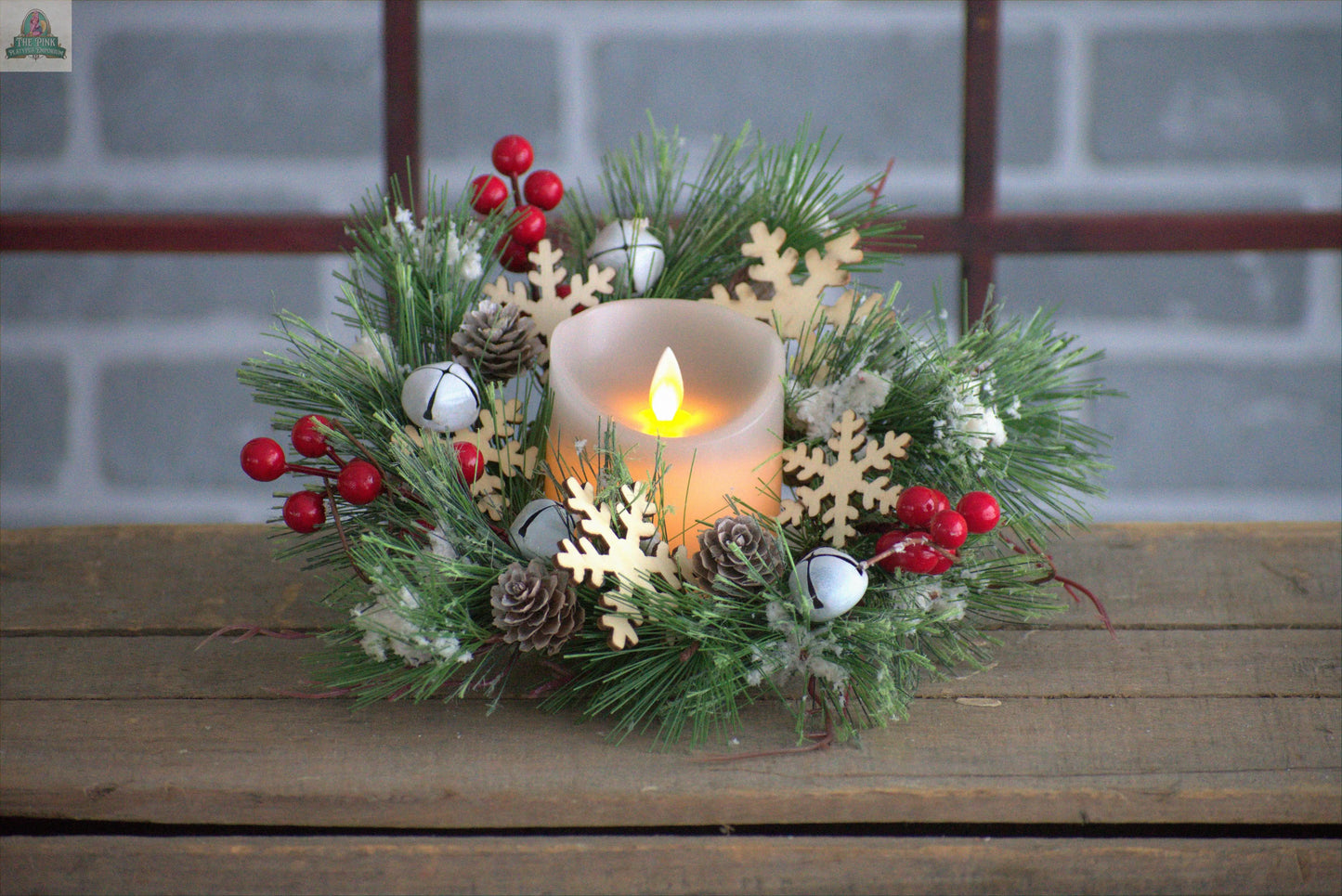 The Pink Platypus Emporium Sleigh Bells 6.5in Candle Ring features faux pine, red berries, pinecones, white jingle bells, and wooden snowflakes for festive decor—perfect on any wooden surface or by a gray brick backdrop.