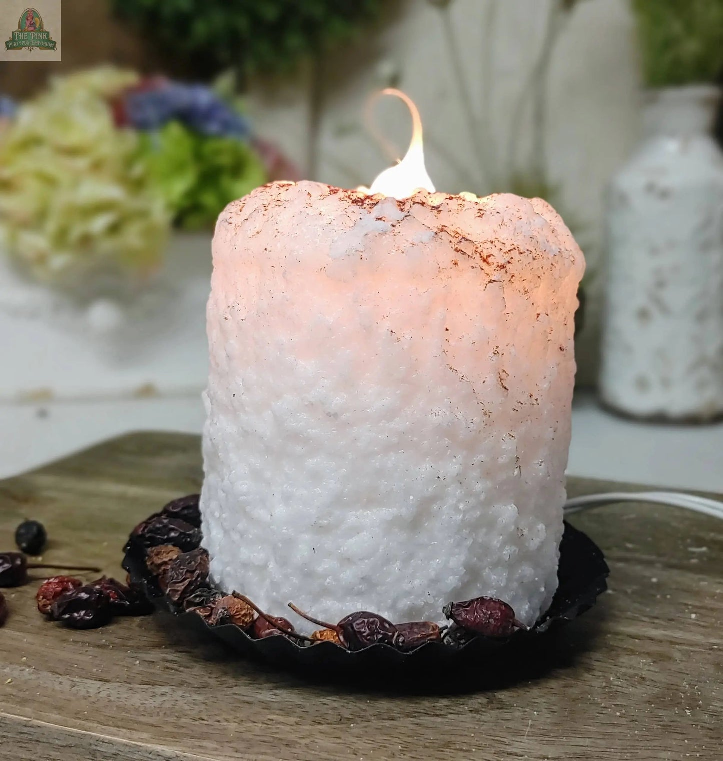 A large white Snickerdoodle Electric Candle glows on a black tray with dried flowers and berries, resting on wood. Blurred flowers and a white vase appear in the background.