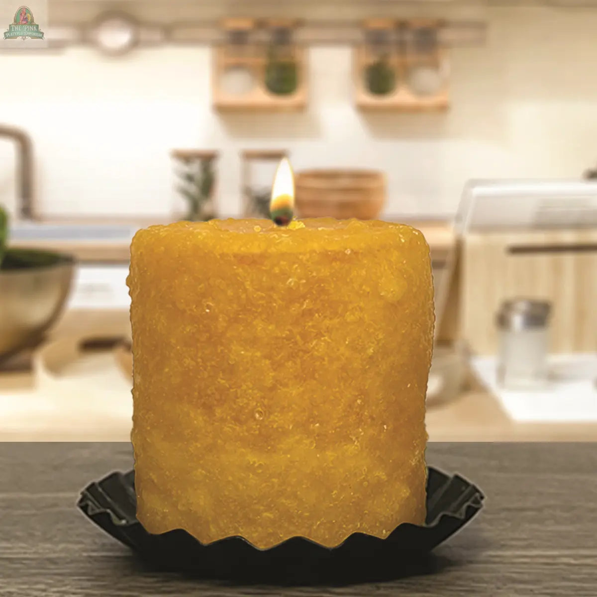 The Spiced Honey Mini Hearth by Pink Platypus Emporium, a textured yellow pillar candle, sits glowing in a black holder on the kitchen counter with shelves and utensils blurred in the background.