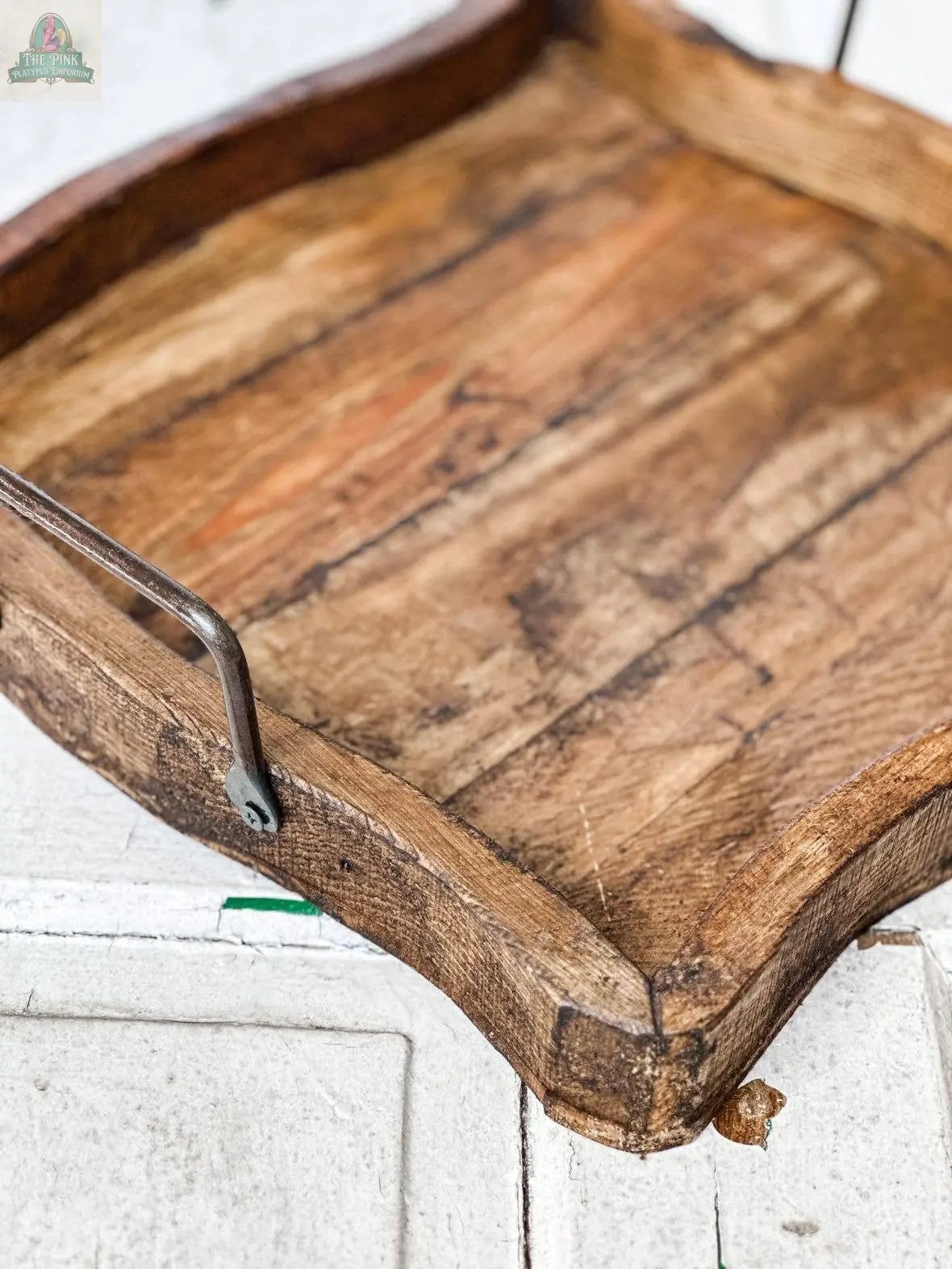 A close-up of the Square Wood Tray, Natural, Handmade - 7.2, featuring curved edges, a metal handle, and weathered grain with worn corners, placed on a white textured surface.