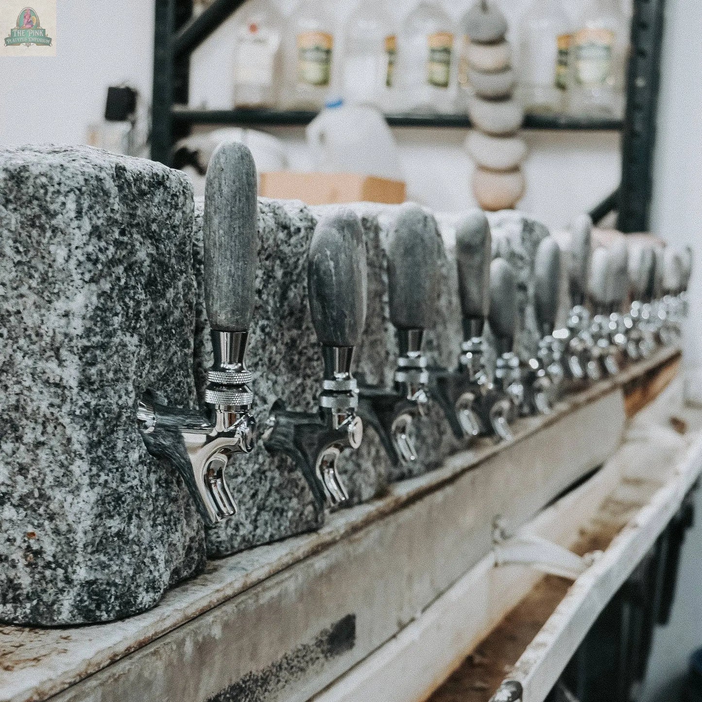 A row of Pink Platypus Emporium Stone Liquor Dispensers with metal spouts and gray handles is arranged on a counter in a workshop, with shelves of barware, bottles, and supplies visible in the background.