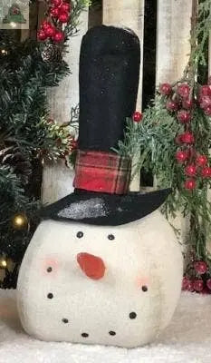 The Tall Black Hat Snowman Head Large features a plush snowman with a carrot nose, black button eyes and mouth, rosy cheeks, and a tall black hat with a red plaid band, sitting on snow amid festive greenery and berries. Size: 12".