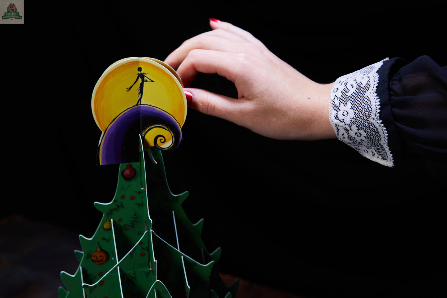 A hand with red nail polish and a lace-trimmed sleeve places a yellow and purple ornament atop the green pop-up tree from The Nightmare Before Christmas: Advent Calendar & Pop-Up Book against a black background.