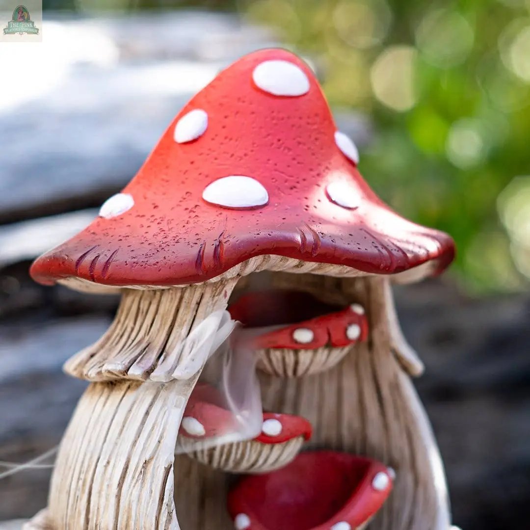 A close-up of the Toadstool Mushroom Backflow Waterfall Incense Burner, featuring a red cap with white spots, inner shelves, and curling smoke. The blurred background shows greenery and wooden planks.