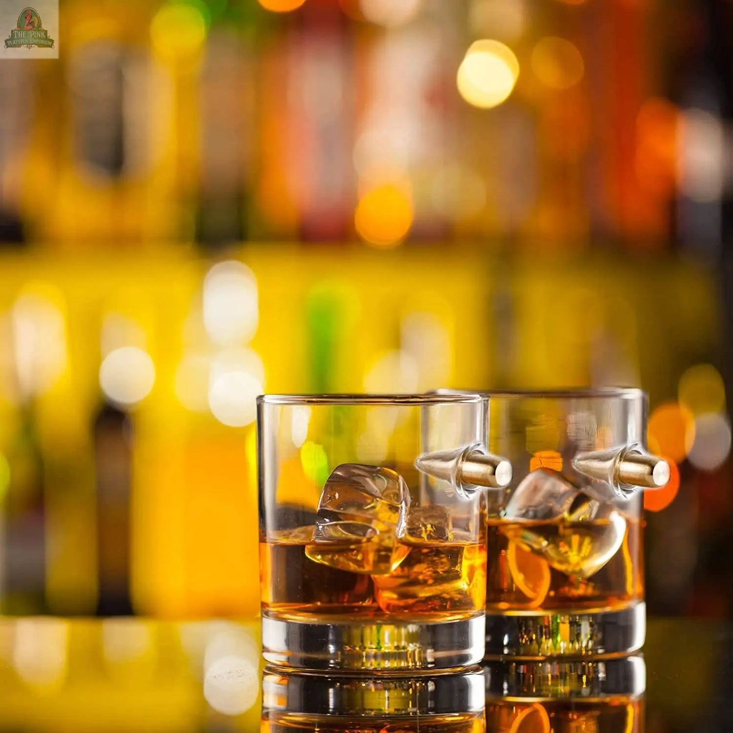 The Pink Platypus Emporium Unique Whiskey Glasses Set of 6 is displayed on a reflective surface, with ice and metal stirrers, backed by blurred bottles and warm yellow lighting.