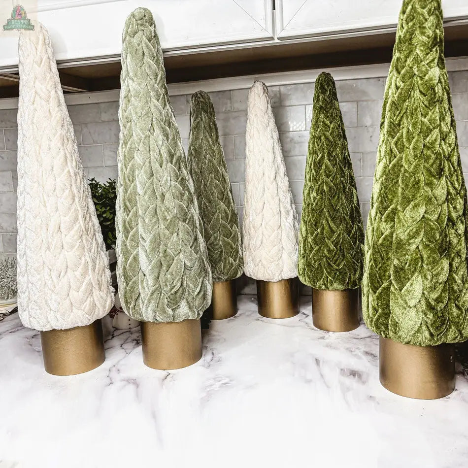 Six Velvet Christmas Trees in green and cream, each with a gold base, are arranged as elegant holiday décor on a marble kitchen countertop with white cabinets and a subway tile backsplash.