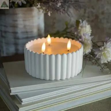 A WHITE CEMENT 3 WICK 3D FLAME CANDLE with a vintage ribbed finish is displayed on stacked white books, its realistic flames glowing warmly as soft-focus flowers and a rustic container accent the background.
