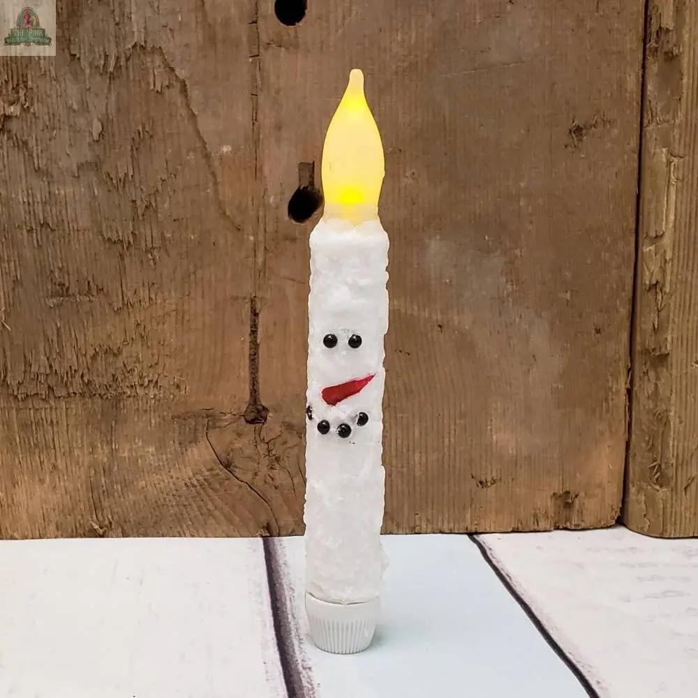 The White Snowman Taper Candle, a battery-operated candle with black eyes, a red mouth, and a textured white body, stands on a white surface with a wooden background.