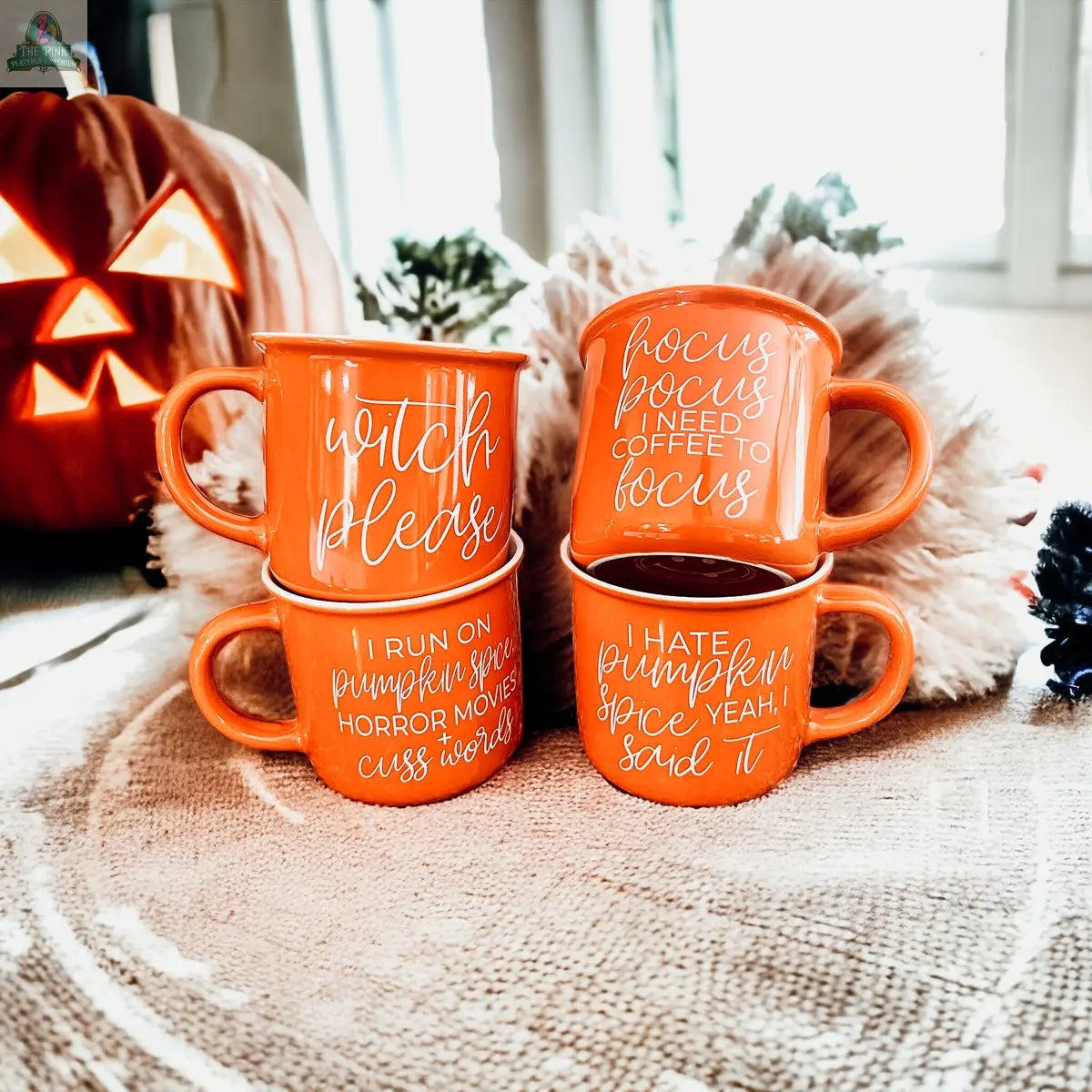 Four orange Witch Please mugs with Halloween phrases are stacked in pairs, with a carved jack-oâ-lantern and autumn decor in the background. Each Halloween coffee mug features playful witch and pumpkin spice sayings.