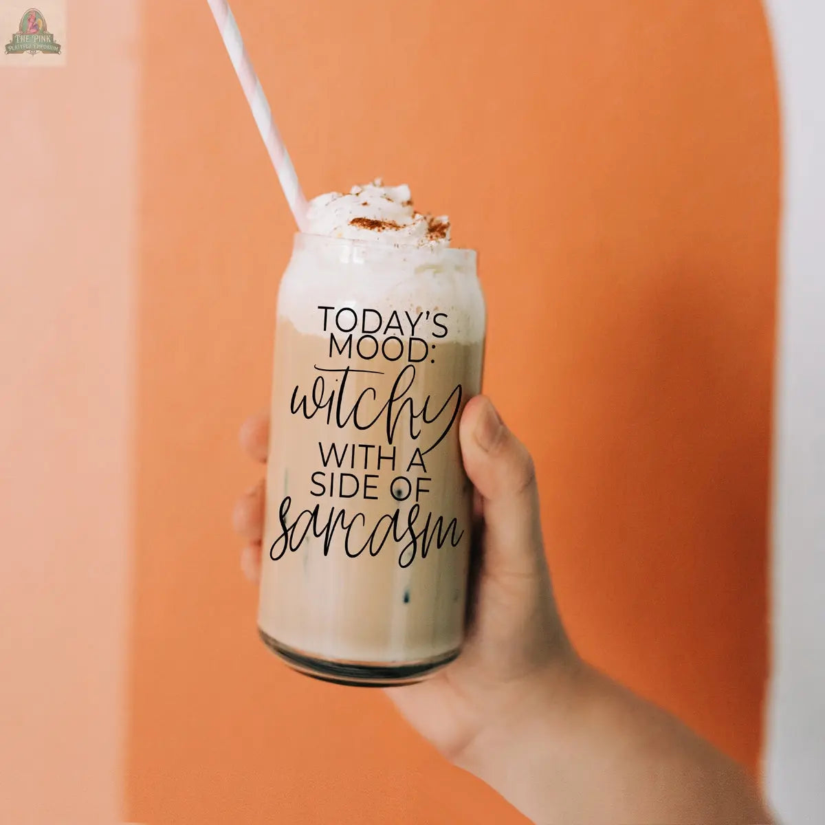 A hand holds the eco-friendly Witchy 20oz tumbler filled with iced coffee, whipped cream, and a straw. The glass reads "Today's mood: witchy with a side of sarcasm" against bold orange and white tones in the background.