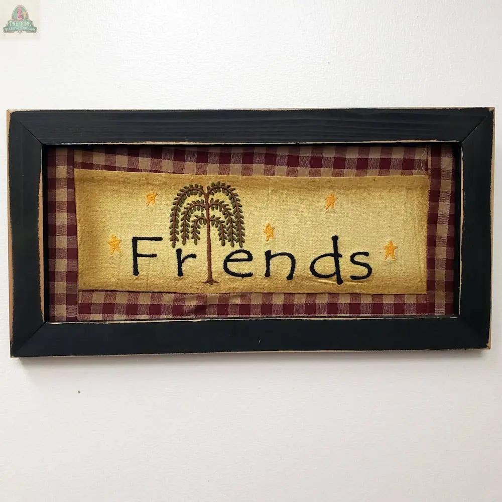 The Wood Framed Friends Stitchery features a painted wood frame, red and tan plaid border, "Friends" stitched in black on yellow with a willow tree for the 'i', and yellow stars—perfect cozy decor for your home.