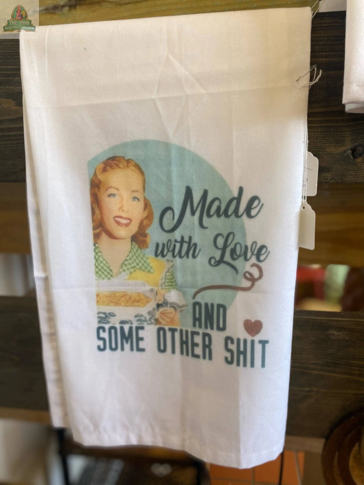The Funny Tea Towel-Made with Love and other shit by Pink Platypus Emporium shows a retro smiling woman with a dish and the phrase "Made with Love and Some Other Shit," perfect for adding quirky charm to your kitchen.