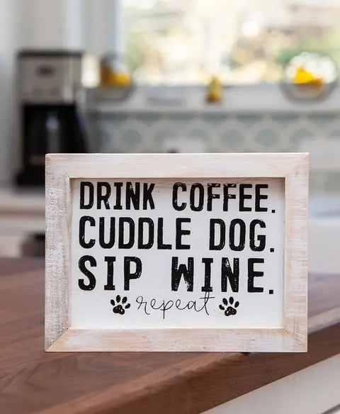 Wooden sign with text 'Drink coffee. Cuddle dog. Sip wine. Repeat.' on a kitchen counter.