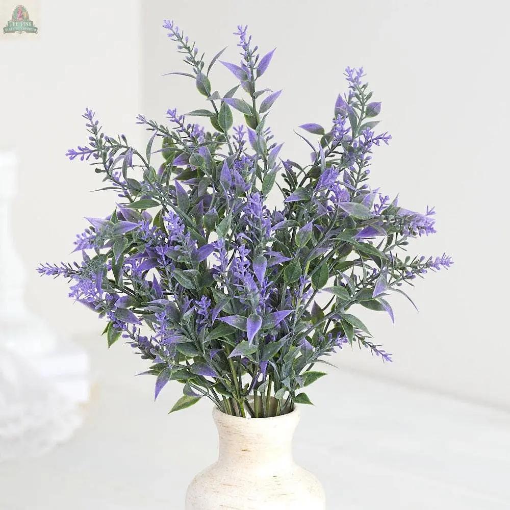 A 13" plastic lavender bush with green leaves sits in a small cream vase, arranged on a light surface against a blurred background.