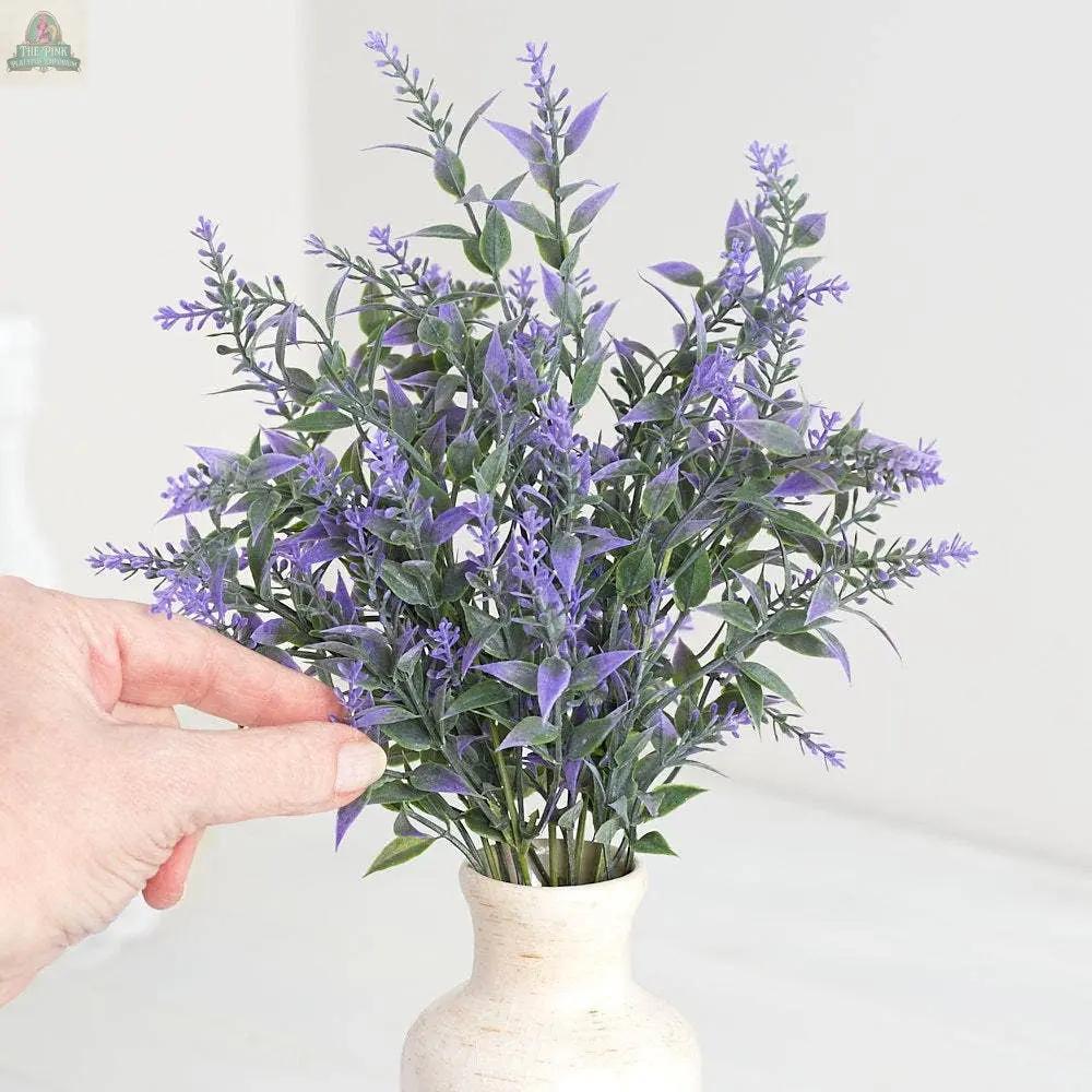 A hand touches a 13" Plastic Lavender Bush with green leaves, arranged in a small white vase on a light surface for a charming decorative floral display.